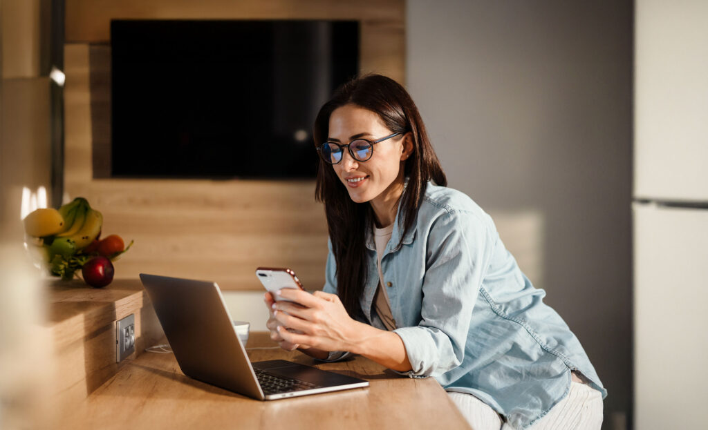 Woman working remotely at a kitchen counter, smiling while using a smartphone and laptop.