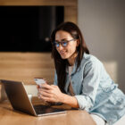 Woman working remotely at a kitchen counter, smiling while using a smartphone and laptop.