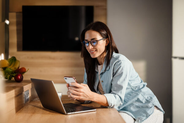 Woman working remotely at a kitchen counter, smiling while using a smartphone and laptop.