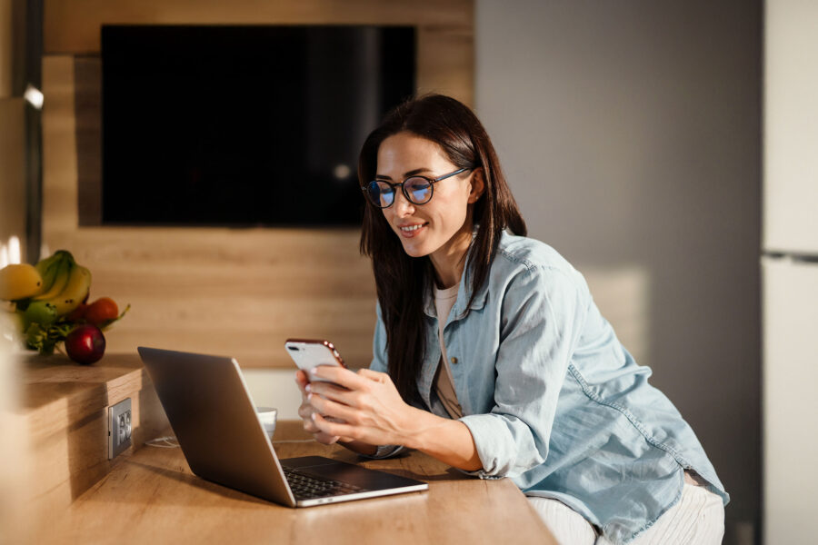 Woman working remotely at a kitchen counter, smiling while using a smartphone and laptop.