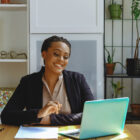A smiling businesswoman sits at a desk working on her laptop in a bright office space, with plants and folders in the background.